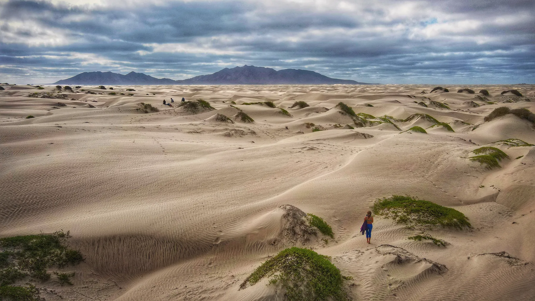 A person walking through a wide dune field beneath cloudy mountains